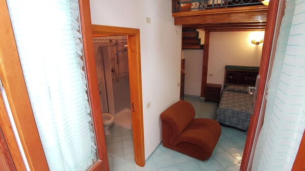 Un couloir étroit avec une porte en bois donnant sur une salle de bain, un petit coin salon et un lit recouvert d’une courtepointe à motifs.