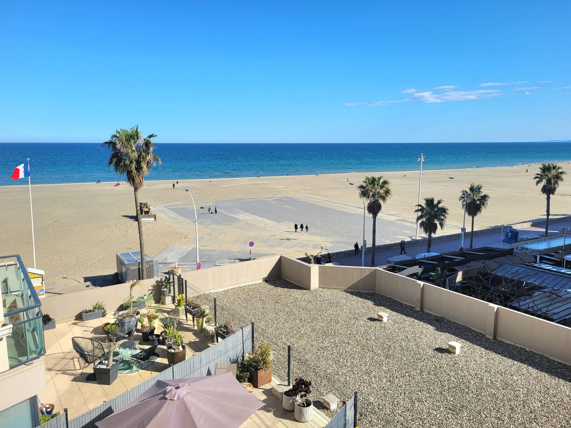 A beach view with palm trees, a flag, and a patio area with outdoor seating.