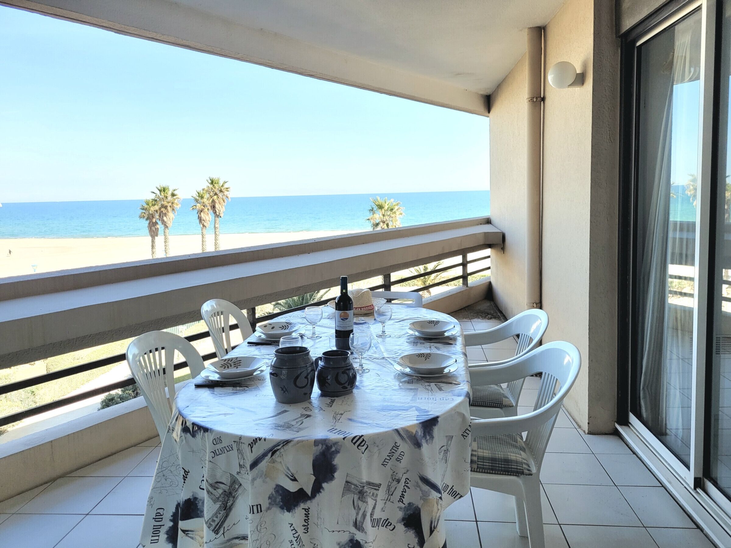 Ein runder Tisch mit Tischdecke, weiße Stühle und eine Weinflasche auf einem Balkon mit Blick auf einen Strand.