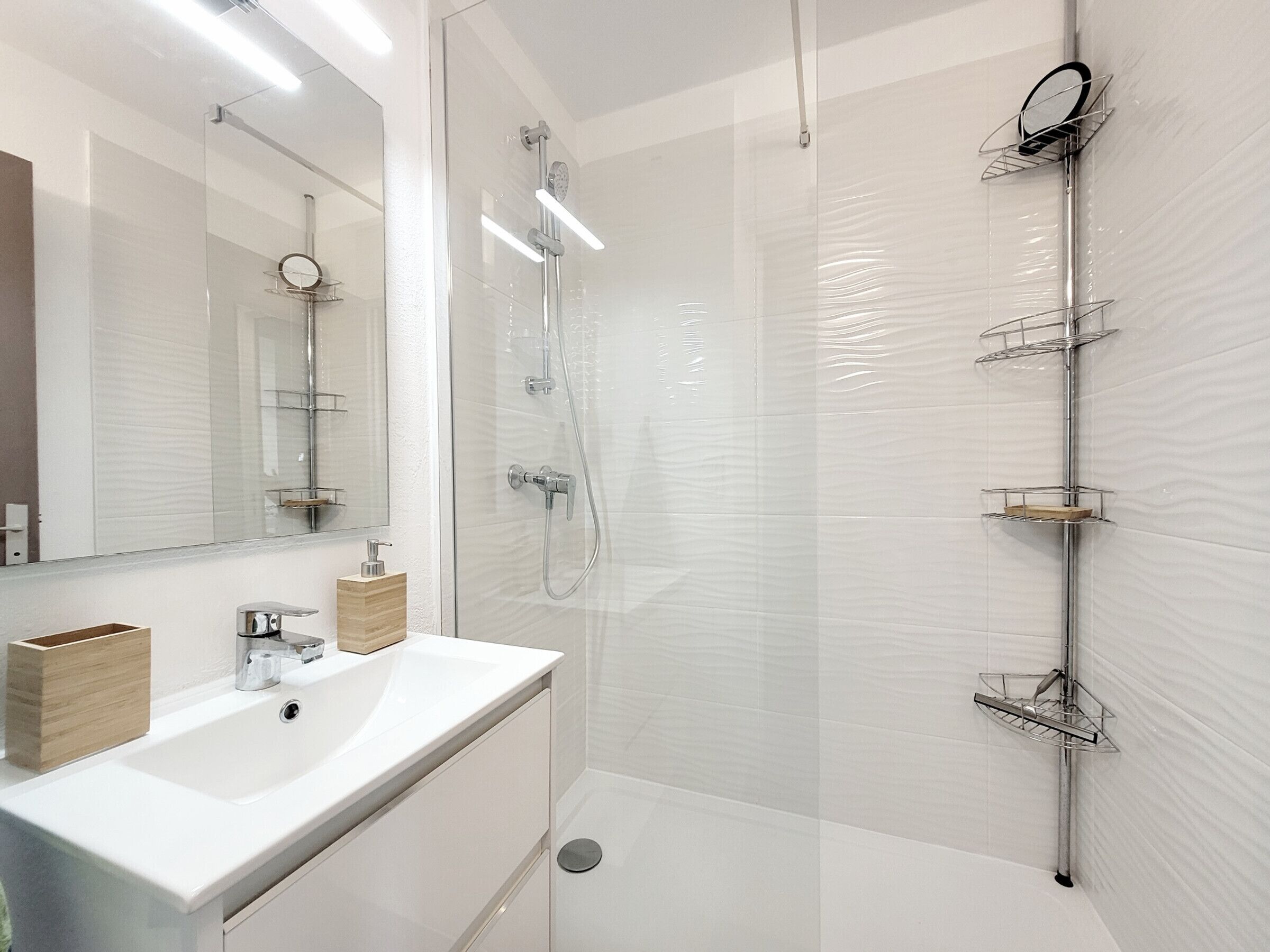 A modern bathroom with a glass-enclosed shower, a white sink with a wooden soap dispenser, and a mirror with a round frame.
