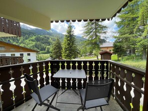Un balcón con una mesa y dos sillas, con vistas a un paisaje montañoso con árboles y edificios.