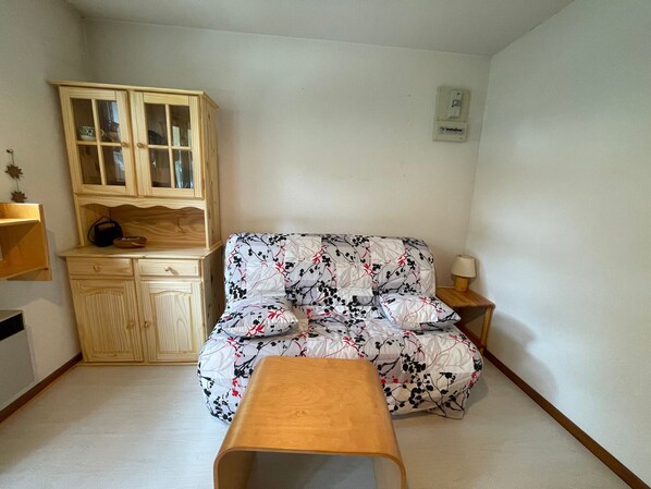 Una habitación pequeña y amoblada con un armario de madera, un sofá con estampado floral, una mesa de madera y una lámpara de noche.