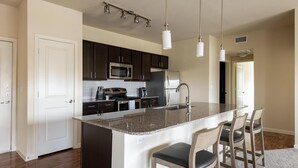 A modern kitchen with dark cabinets, stainless steel appliances, a granite countertop, and pendant lighting. - Landing | Chic 3BD, Clubhouse, Pool (Austin)