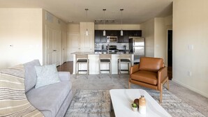 A modern living room with a grey sofa, a brown leather chair, and a white coffee table with decorative items. A kitchen with dark cabinets and stainless steel appliances is visible in the background. - Landing | Chic 3BD, Clubhouse, Pool (Austin)