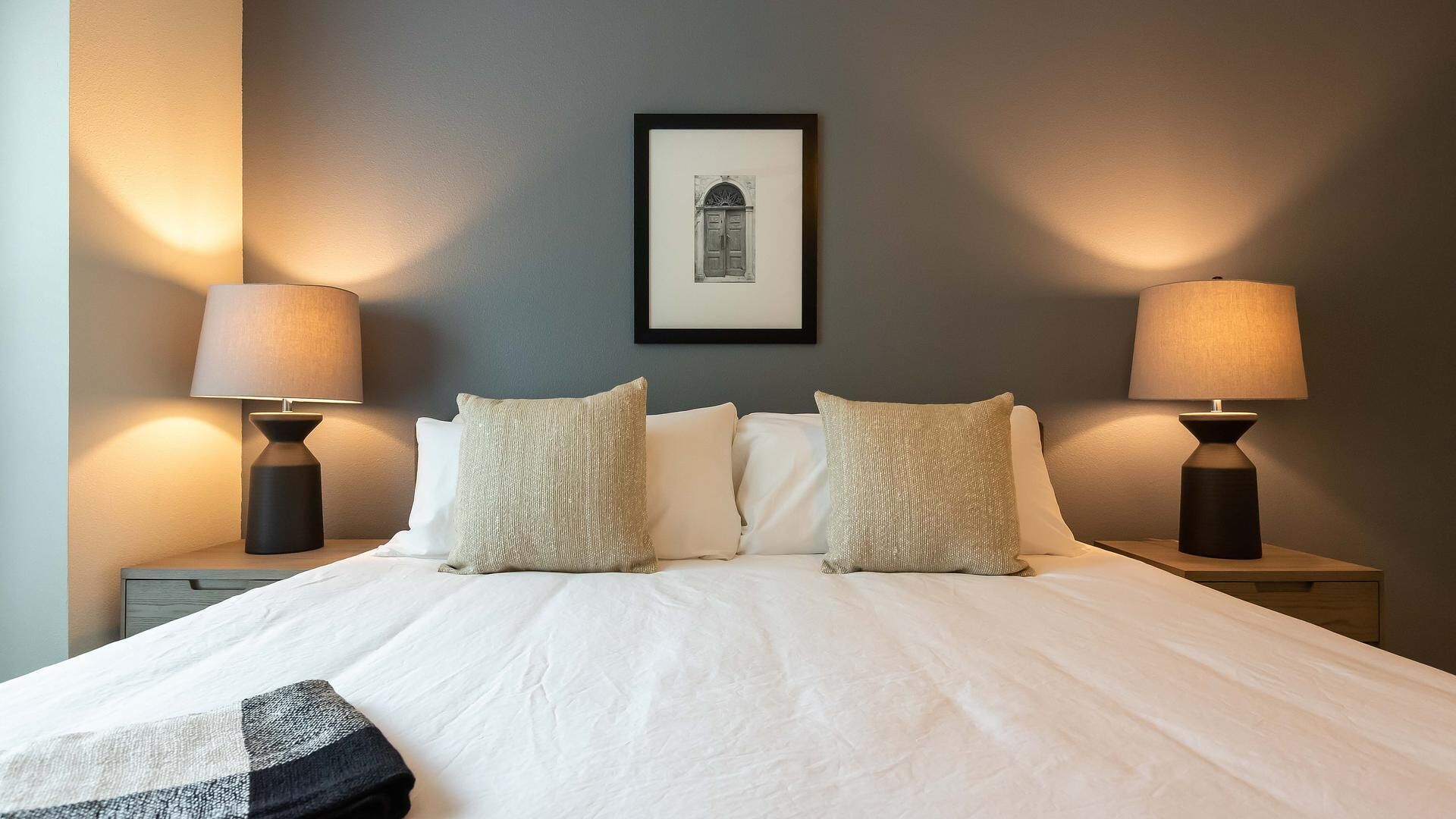 A neatly made bed with white linens and two decorative pillows, flanked by two bedside tables with lamps, in a room with a framed picture on the wall.