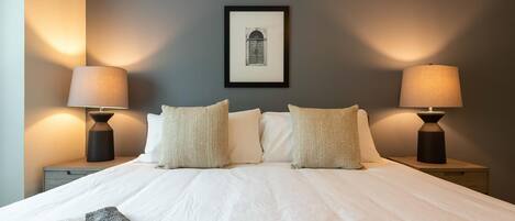 A neatly made bed with white linens and two decorative pillows, flanked by two bedside tables with lamps, in a room with a framed picture on the wall.