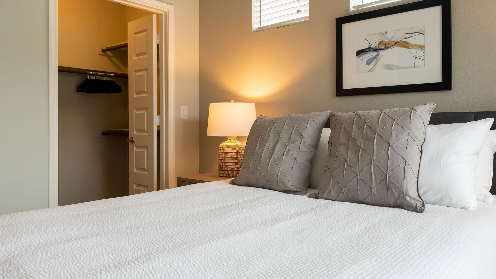 A hotel room with a bed, a nightstand with a lamp, a framed abstract painting, and a closet with hanging clothes.