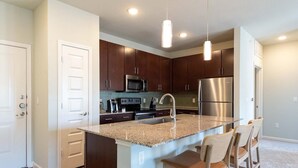 A modern kitchen with dark wood cabinets, stainless steel appliances, and a granite countertop island with bar stools. - Landing | Amazing 2BD, Gym, Clubhouse (Austin)