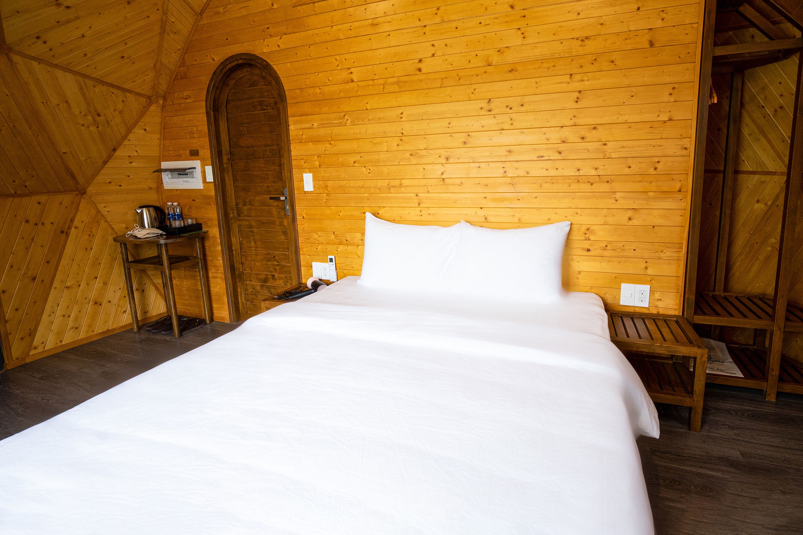 A cozy wooden room with a bed, a small table with a kettle, and a wooden bench.