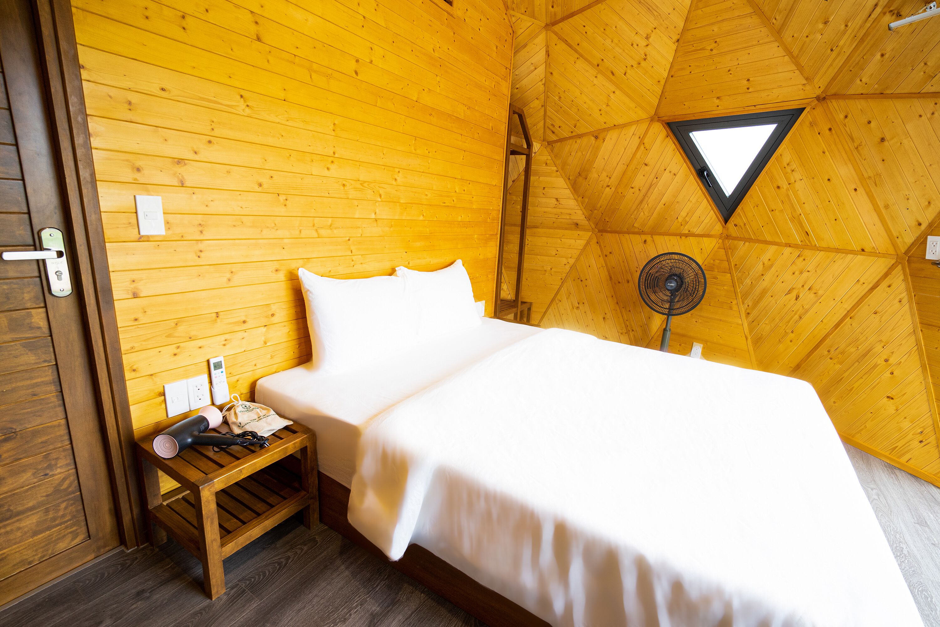 A cozy room with a bed, a small wooden table, and a fan.