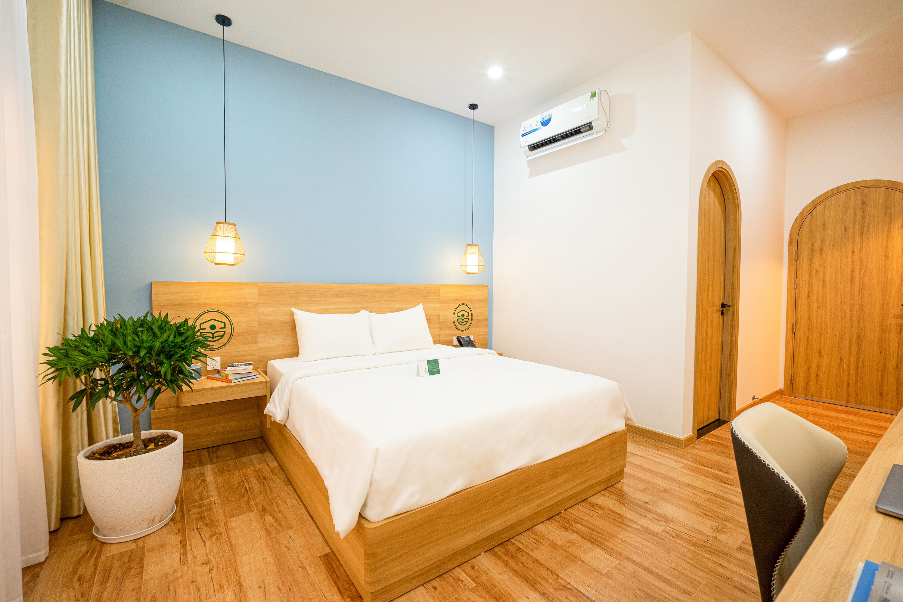 Habitación de hotel con cama, mesita de noche, lámpara, planta en maceta y armario de madera.