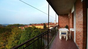Een balkon met een tafel en stoelen, met uitzicht op een bebost gebied en gebouwen in de verte.