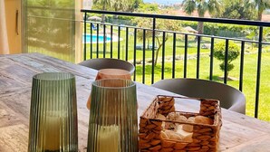 Una mesa de madera con dos candelabros de vidrio y una cesta tejida en un balcón con vista a una piscina y palmeras.