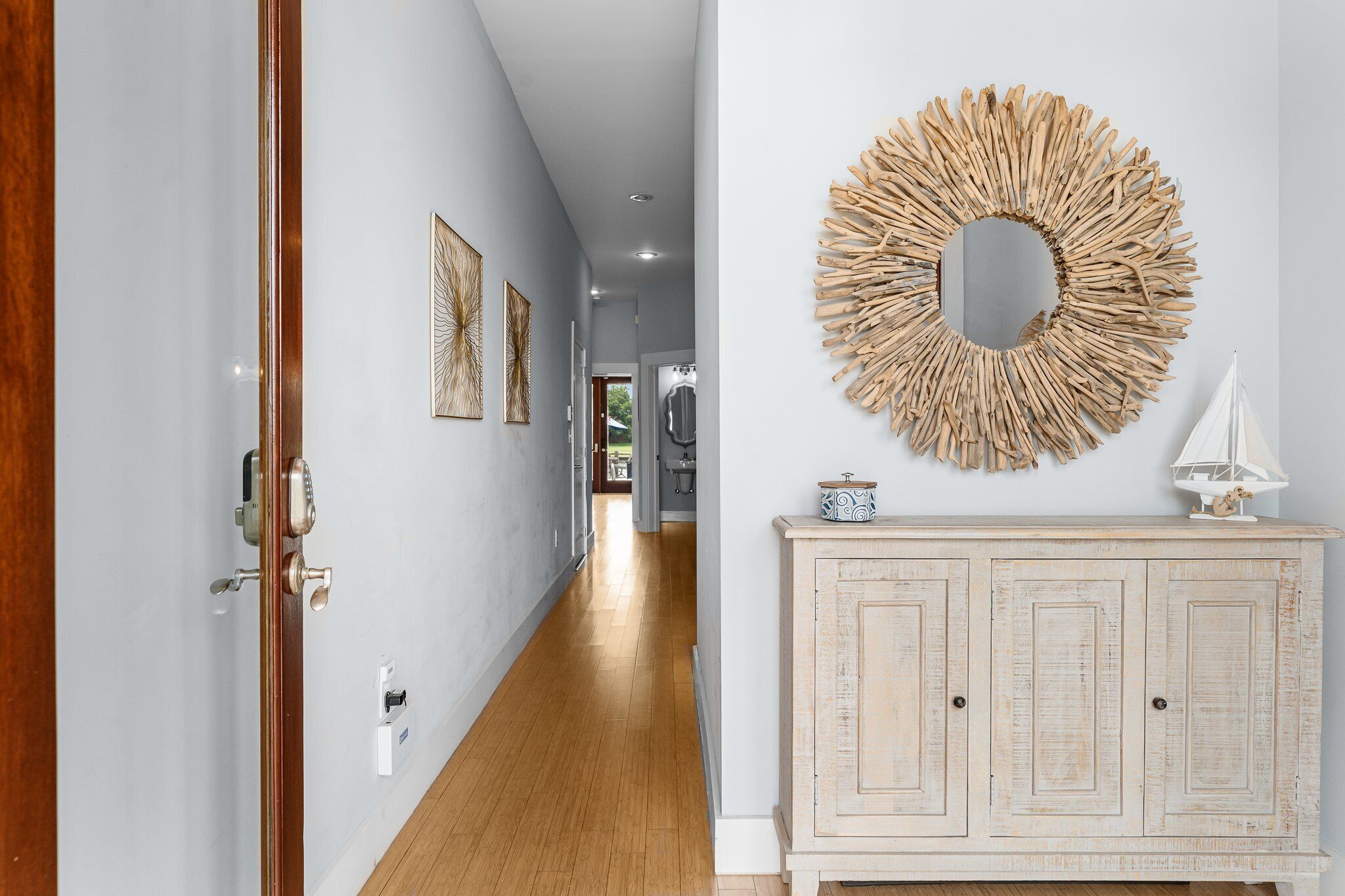Un pasillo con suelo de madera, una pared blanca con un espejo circular y un armario blanco decorado con un velero.