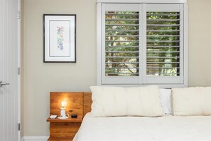 A bedroom with a bed, a nightstand with a lamp, a framed picture on the wall, and a window with blinds. - Luxe Cape May Point Home near Beach +Peloton (Cape May)