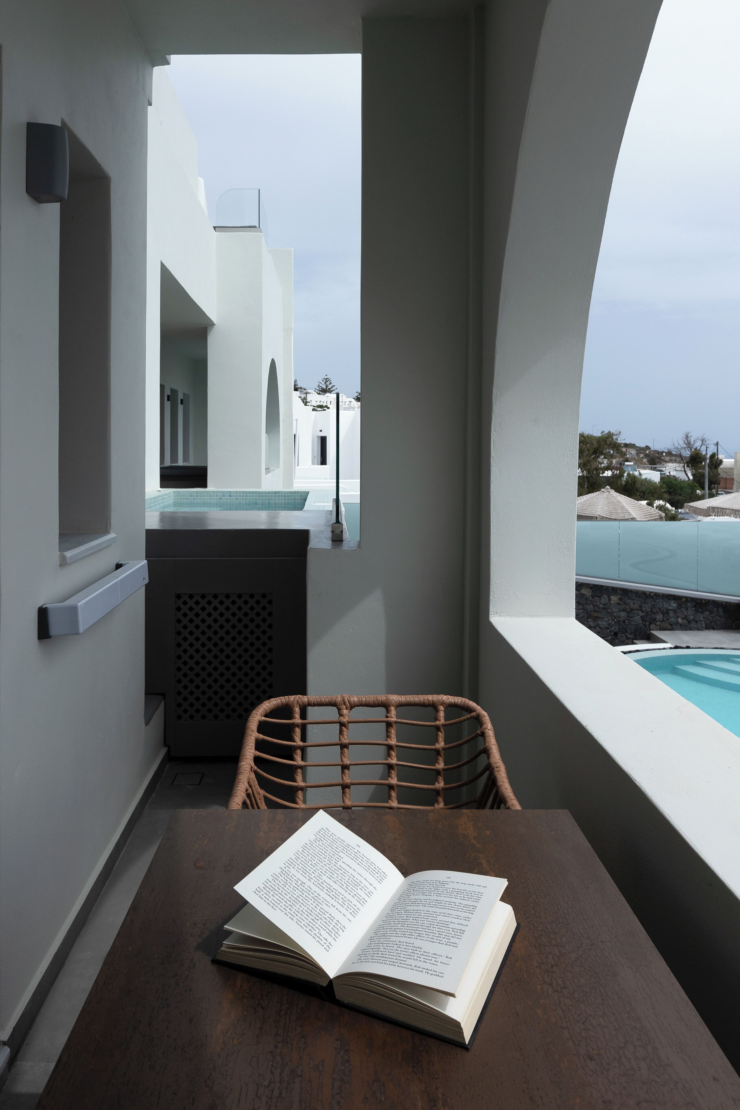 A modern room with a view of a pool and buildings, a wooden table with an open book, and a wicker chair.