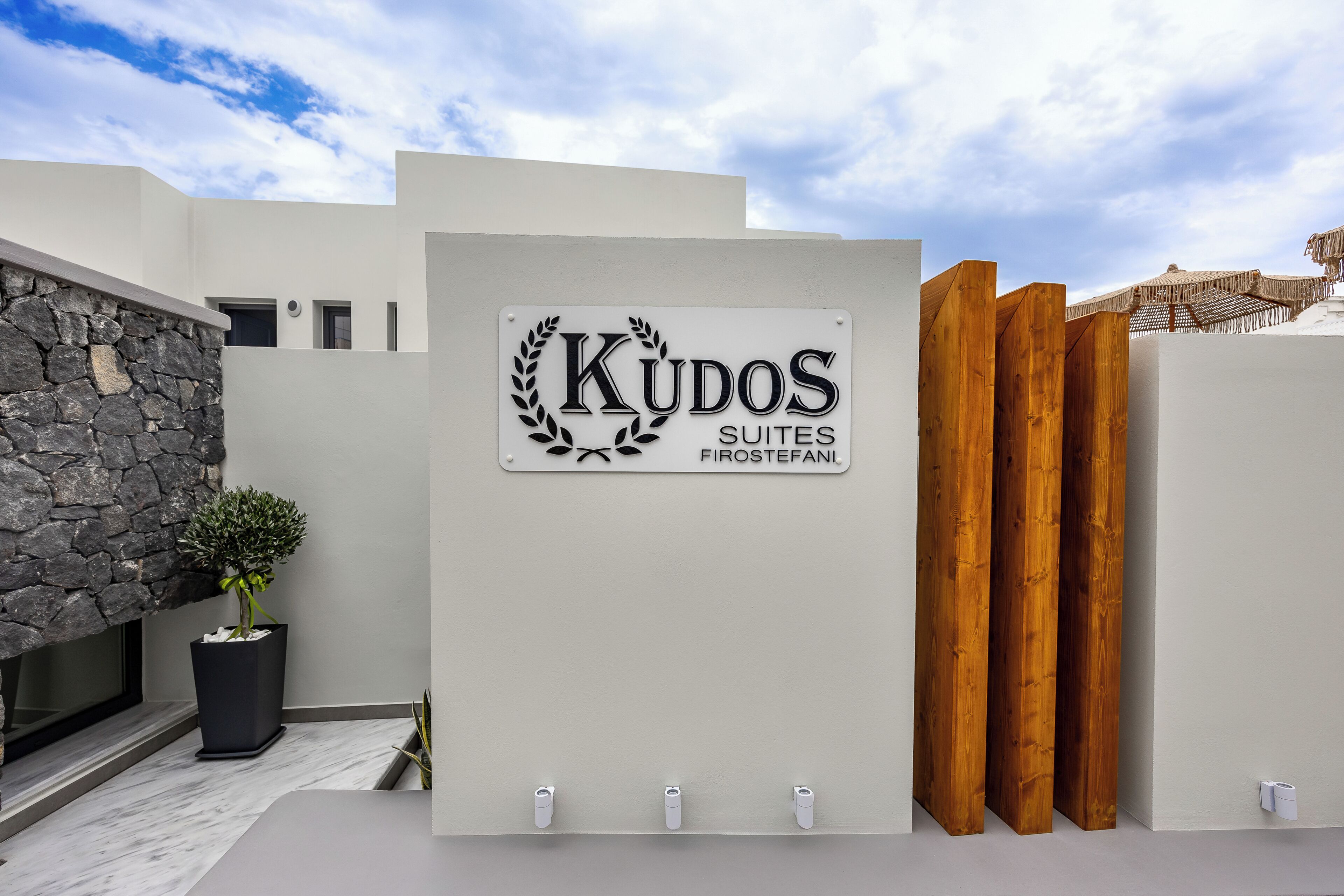 A sign for 'Kudos Suites' with a modern architectural design, a stone wall, and a potted plant.