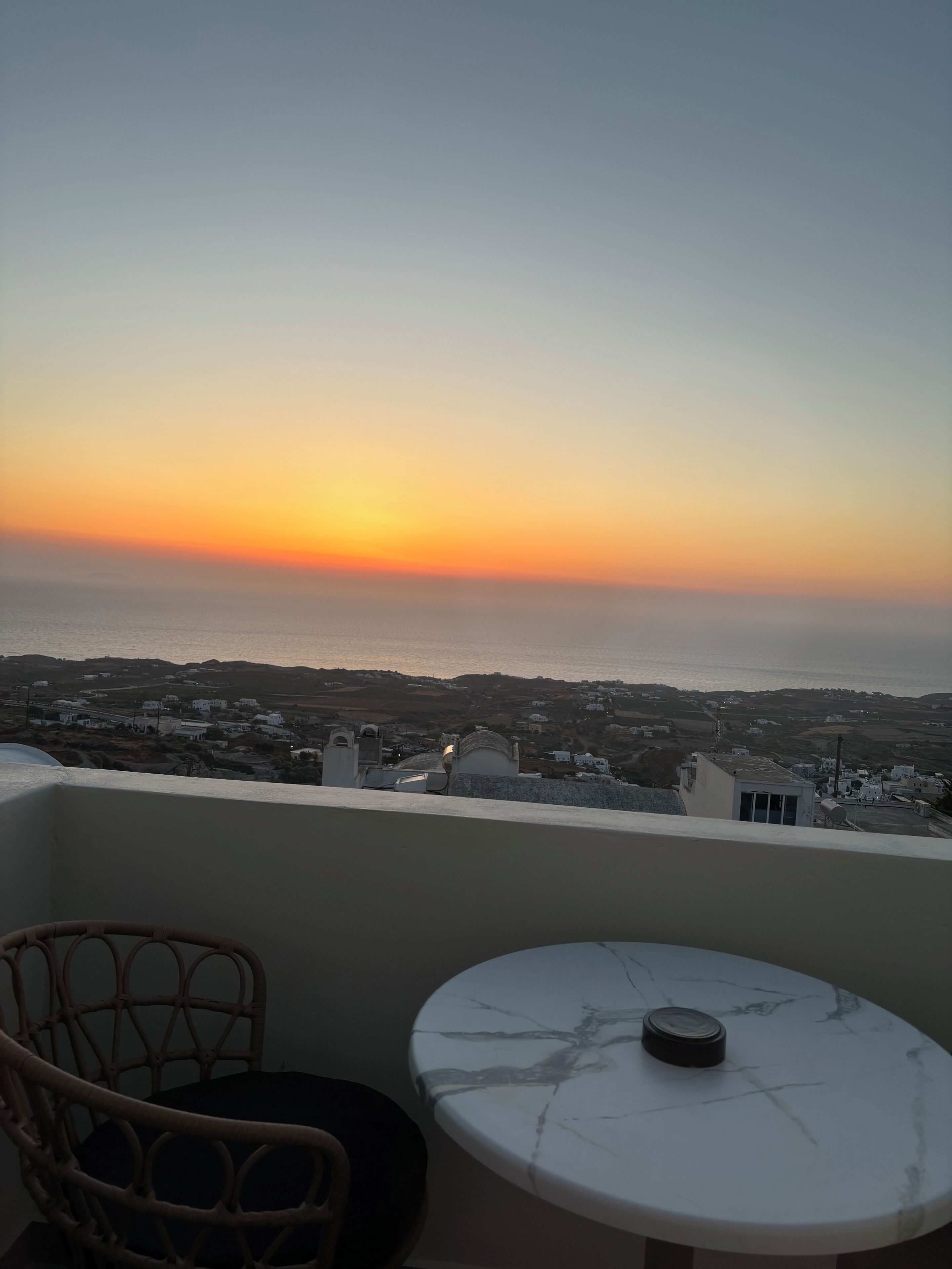 A balcony with a view of the sea at sunset, a round table, and wicker furniture.