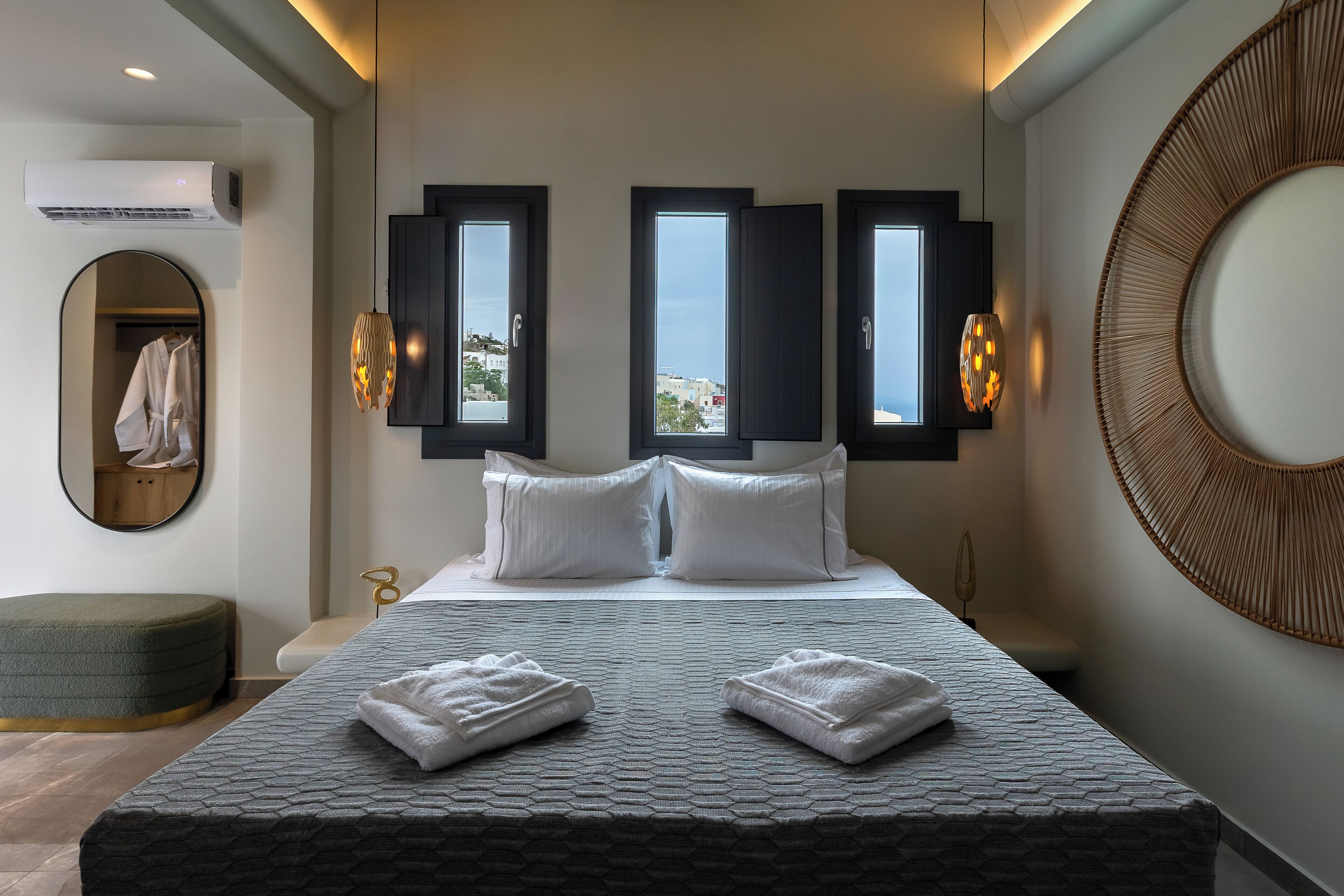 A modern hotel room with a large bed, two windows with shutters, a round mirror, and a wall-mounted air conditioner.