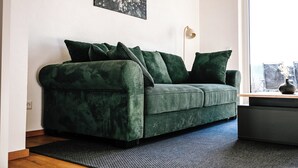 A green sofa with a textured fabric, a dark rug, a wall-mounted picture, and a window with a view. - GardenOasis: Exclusive Seehalde apartment (Schluchsee)