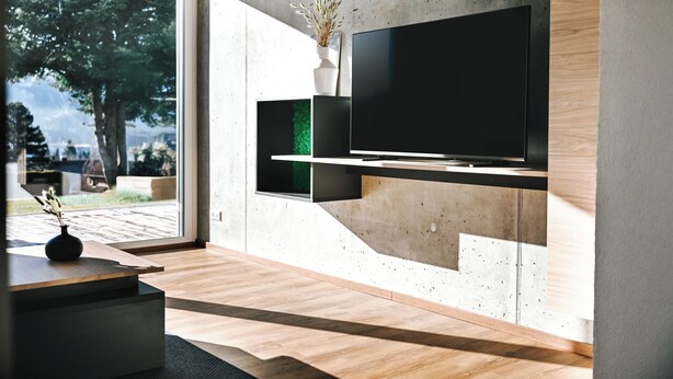 A modern living room with a large flat-screen TV mounted on a concrete wall, wooden flooring, and a view of a tree and outdoor area. - GardenOasis: Exclusive Seehalde apartment (Schluchsee)