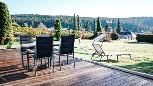 A wooden deck with outdoor furniture, including chairs and a table, overlooking a grassy area with a view of a forested hill and a windmill. - GardenOasis: Exclusive Seehalde apartment (Schluchsee)