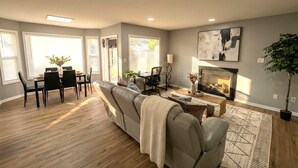 A living room with a sofa, a dining table with chairs, a fireplace, and a large window. - Renovated 3BR — King Suite, Laundry, Parking (New Castle)