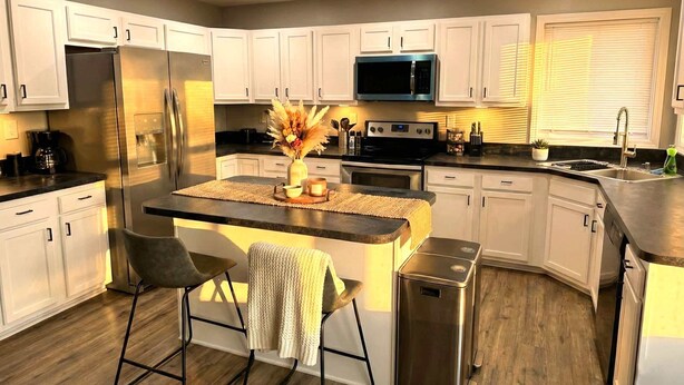A modern kitchen with white cabinets, stainless steel appliances, a central island with bar stools, and a microwave above the island. - Renovated 3BR — King Suite, Laundry, Parking (New Castle)