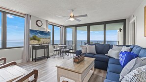 Ein großzügiges Wohnzimmer mit einer großen blauen Couch, einem Holztisch und einem Essbereich mit Tisch und Stühlen. Ein Deckenventilator, ein Gemälde an der Wand und ein Blick auf das Meer durch große Fenster sind ebenfalls vorhanden.