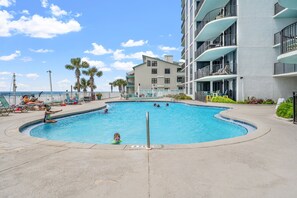 A swimming pool surrounded by lounge chairs and palm trees, with a building in the background. - Watercrest 1502 - Walkin on Sunshine (Panama City Beach)