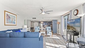 Ein modernes Wohnzimmer mit einem blauen Sofa, einem Essbereich mit Tisch und Stühlen, einer Küche im Hintergrund und einem großen Fenster mit Blick auf den Strand.