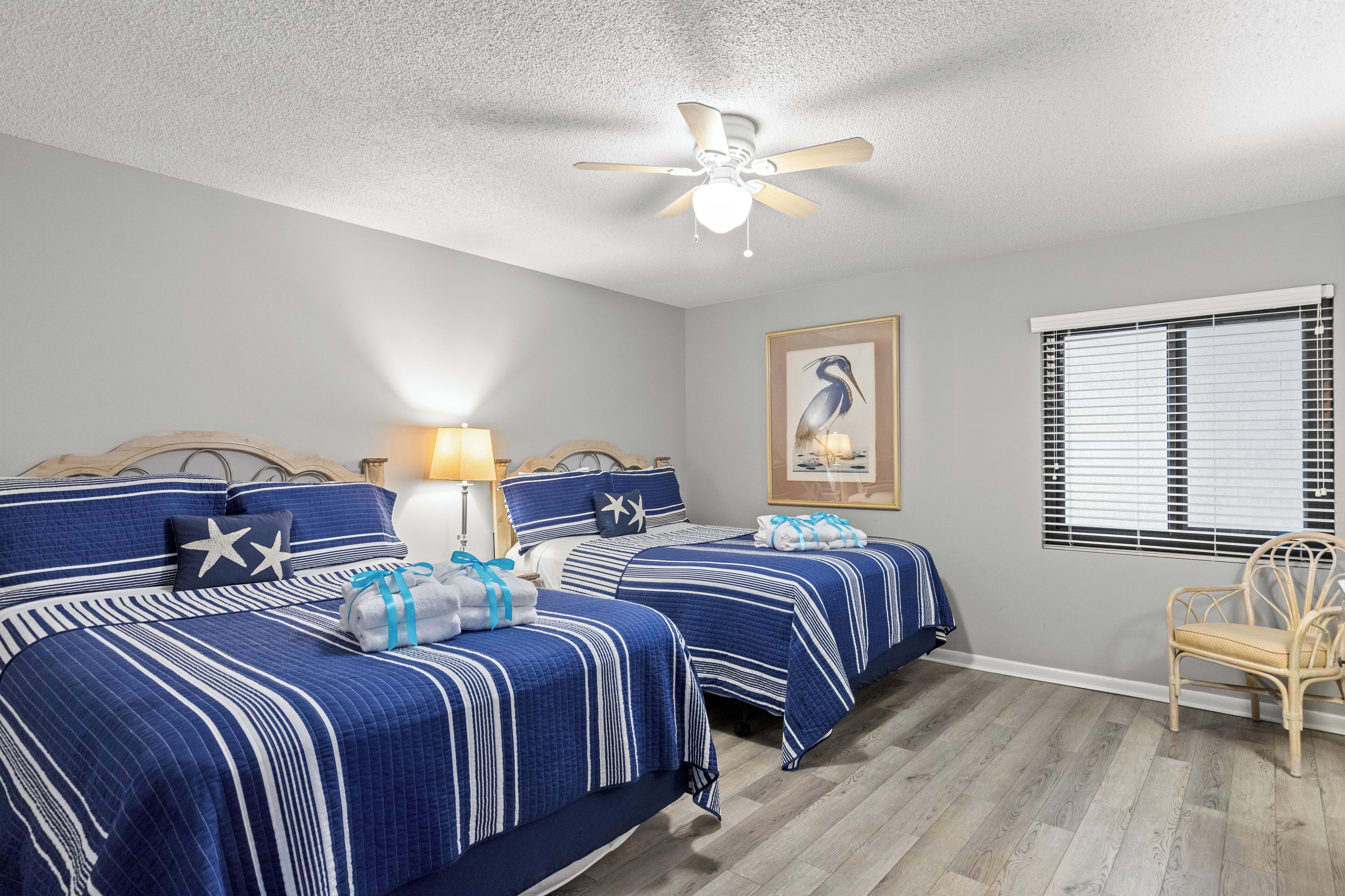 A bedroom with two beds, a ceiling fan, a window with blinds, and a framed picture on the wall.