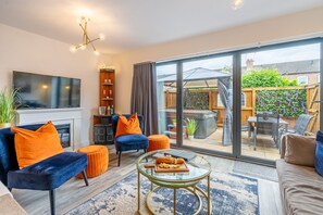 A modern living room with a TV, comfortable seating, a glass coffee table with snacks, and a view of the backyard through large sliding doors. - The Ebor House - sleeps 7 guests in 3 bedrooms with hot tub (York)