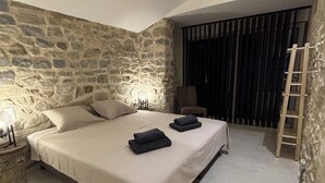 Ein Schlafzimmer mit Steinwand, einem Bett mit zwei Kissen, zwei gefalteten Handtüchern, einem Sessel, einer Leiter und einem Fenster mit Jalousien.