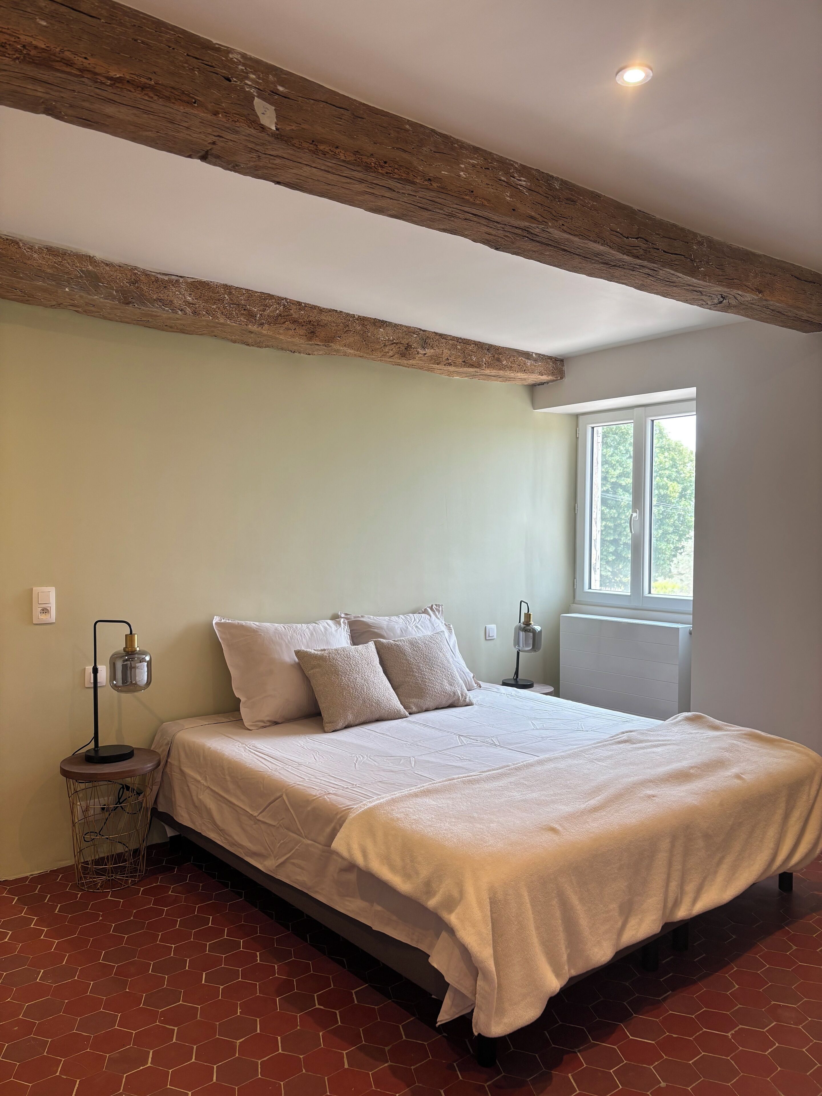Une chambre avec un grand lit, une table de chevet avec une lampe, une fenêtre donnant sur un paysage verdoyant et des poutres apparentes en bois au plafond.