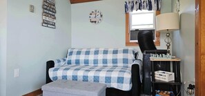 Una habitación con un sofá a cuadros, un pequeño otoman, un reloj de pared, una lámpara y una estantería con objetos.