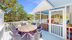 A covered outdoor deck with a round wooden table and wicker chairs, surrounded by white railings and overlooking a lush garden.