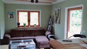 A living room with a brown sofa, a glass coffee table, a wooden dining table, and a window with blinds. - Gemütliches Ferienhaus im Grünen (Michendorf)