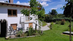 A white house with a wooden staircase leading to a deck, a small outdoor seating area with a table and chairs, and a well-maintained garden with a variety of plants and a clear sky. - Gemütliches Ferienhaus im Grünen (Michendorf)