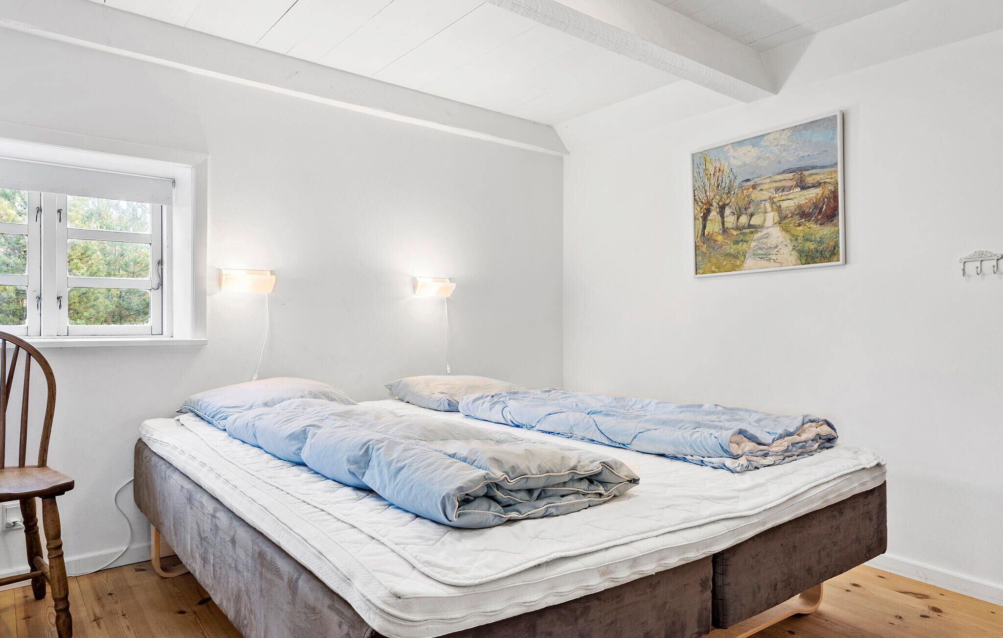 A bedroom with two beds, a wooden chair, a window with white shutters, and a painting on the wall.