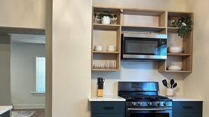 Una cocina moderna con estufa, horno y microondas integrados, estantes de madera con vajilla, y un elegante accesorio de iluminación.