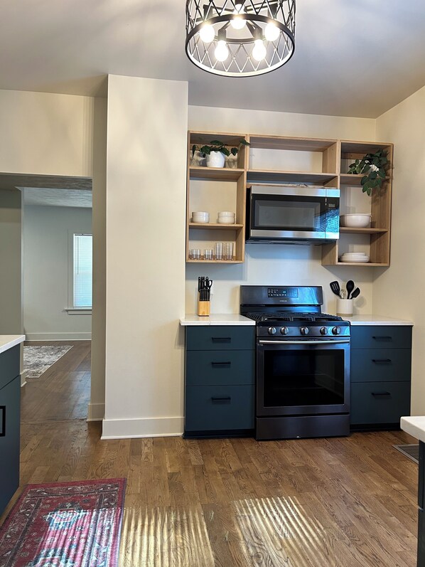 Une cuisine moderne dotée d’une cuisinière, d’un four et d’un four à micro-ondes encastrés, d’étagères en bois où sont disposées de la vaisselle, et d’un luminaire décoratif.