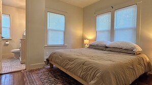 Un dormitorio con cama, ventilador de techo, dos ventanas con persianas, una mesita de noche con lámpara y una alfombra estampada.
