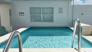 A swimming pool with a metal ladder, a tiled deck, and a building with a window in the background.