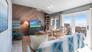 A spacious living room with a large TV, comfortable seating, a wooden herringbone wall, and a balcony with a view. - Lost Colony is a beach front complex with a Community pool and pedestrian boardw (Port Aransas)