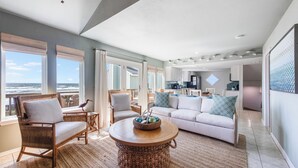 A spacious living area with a view of the ocean, a round wooden coffee table, a white sofa, and wicker chairs. - Lost Colony is a beach front complex with a Community pool and pedestrian boardw (Port Aransas)