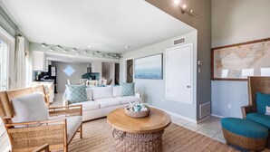 A spacious living room with a large round coffee table, wicker furniture, and a view into the kitchen. - Lost Colony is a beach front complex with a Community pool and pedestrian boardw (Port Aransas)