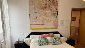 Una cama bien hecha con almohadas florales, un cuadro abstracto grande en la pared y una mesita de noche con una lámpara.