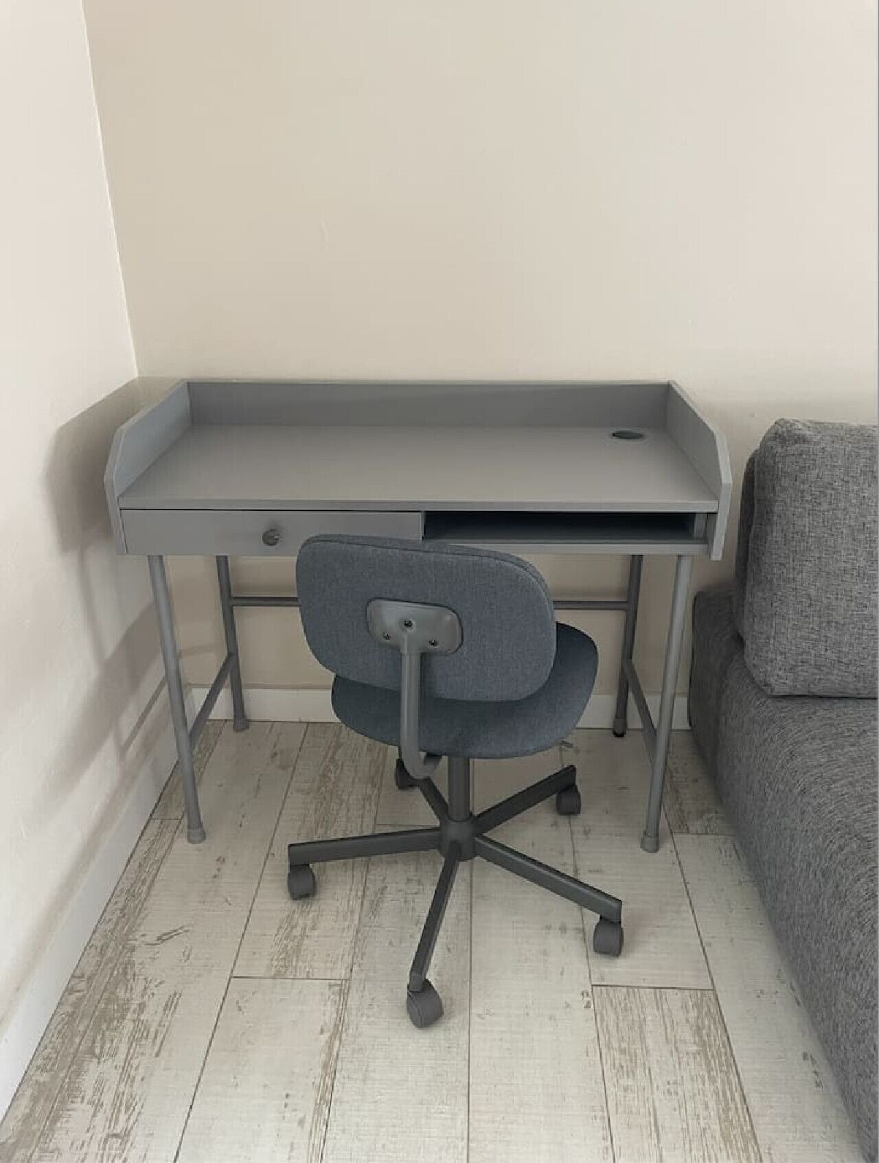 A corner desk with a grey office chair in a room with light wooden flooring and a grey sofa.