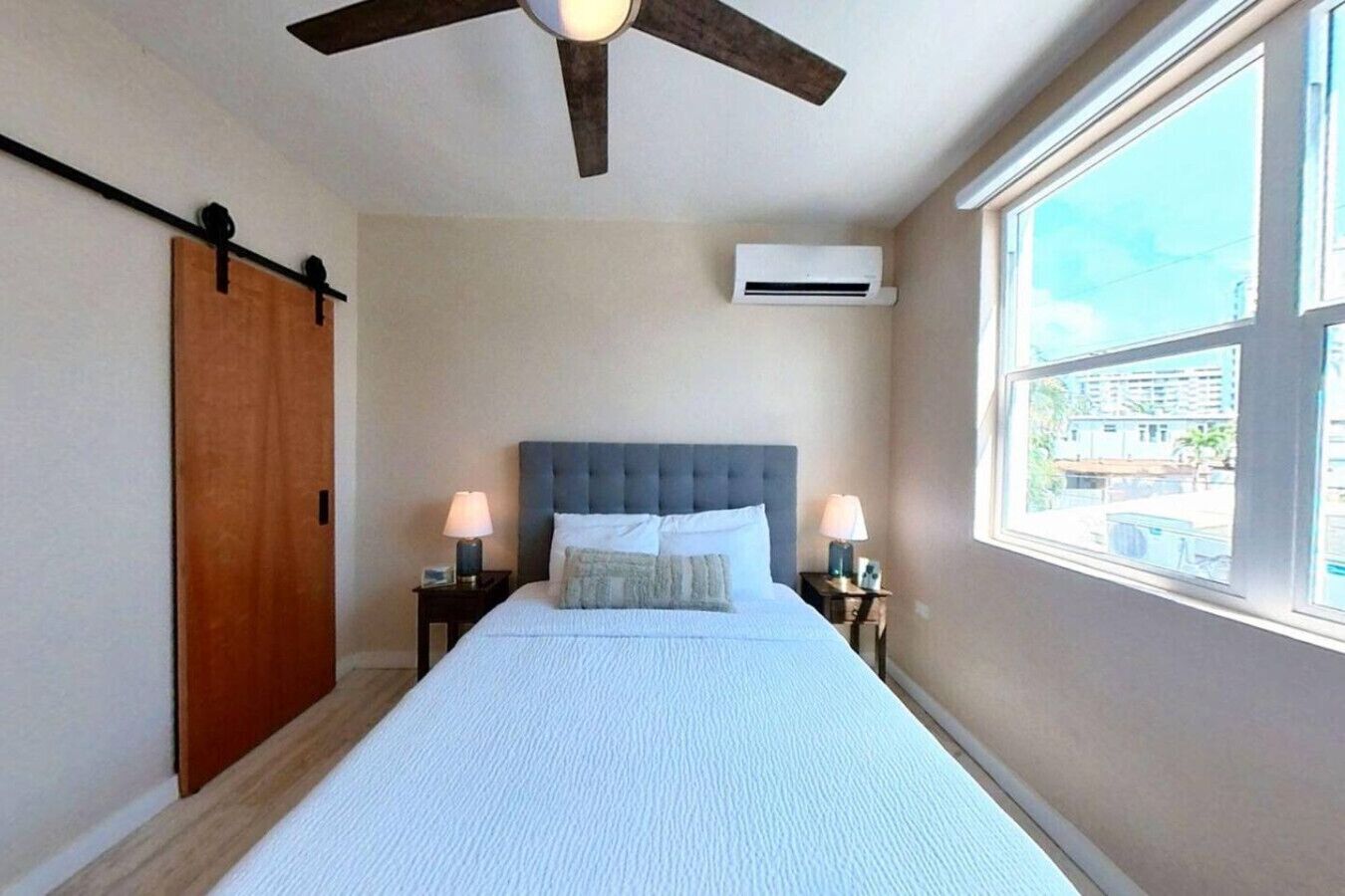 Un dormitorio con una cama grande, un ventilador de techo, un equipo de aire acondicionado y una ventana con vistas a edificios.